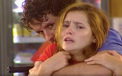 Um ator (nome não divulgado) e Deborah Secco em cena de Laços de Família: ele abraça a atriz por trás e está com uma arma em uma das mãos
