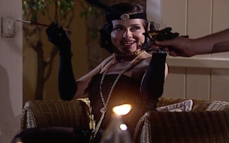 A atriz Soraya Ravenle com uma roupa de melindrosa preta, segura um cigarro com a mão esquerda e uma taça de espumante com a mão direita em cena de Laços de Família