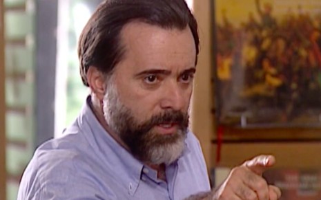 O ator Tony Ramos aponta o dedo para a direita em uma livraria caracterizado como o Miguel em cena de Laços de Família