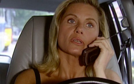A atriz Vera Fischer dirige um carro enquanto fala no celular caracterizada como a Helena em cena de Laços de Família