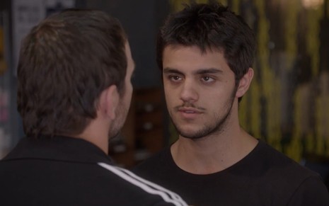Felipe Simas grava de camiseta preta e expressão séria com Marcelo Faria, de costas, em Malhação Sonhos