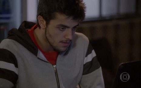 Felipe Simas grava cena de Malhação com casaco cinza com a manga listrada, cabelo arrepiado e expressão séria olhando para o computador como Cobra