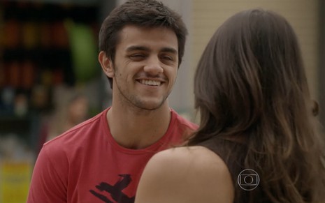 Felipe Simas grava de camiseta vermelha e sorriso no rosto olhando para Anaju Dorigon, de costas na foto