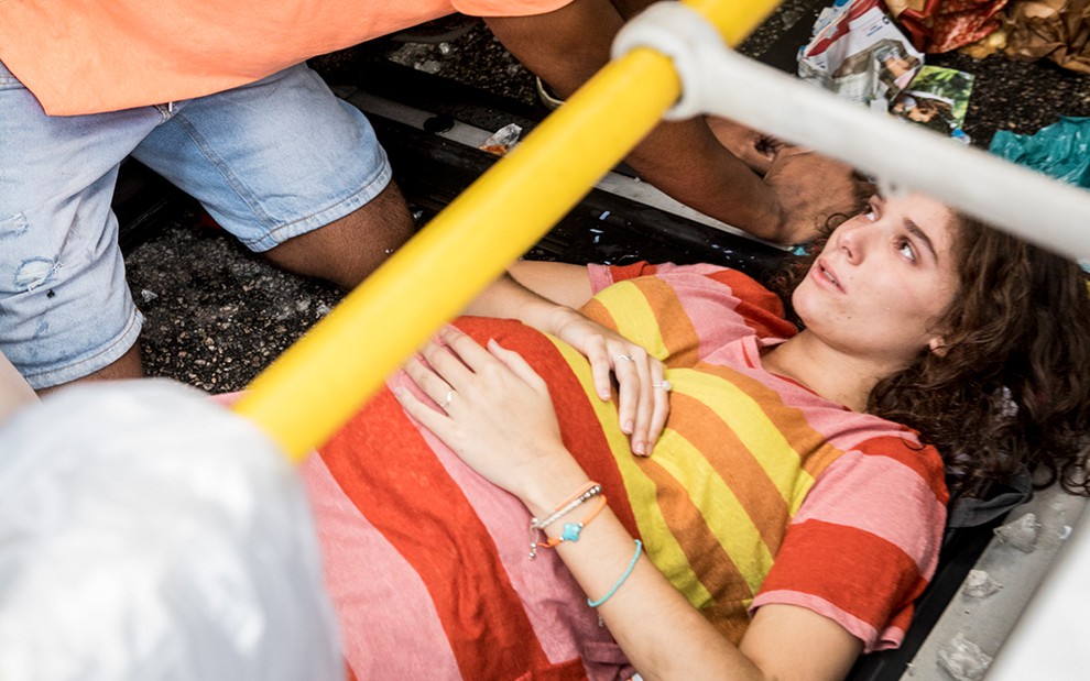 A atriz Giulia Bertolli, com um vestido listrado, deitada em meio às ferragens de um ônibus, machucada e suja, caracterizada como a Meg de Malhação: Toda Forma de Amar