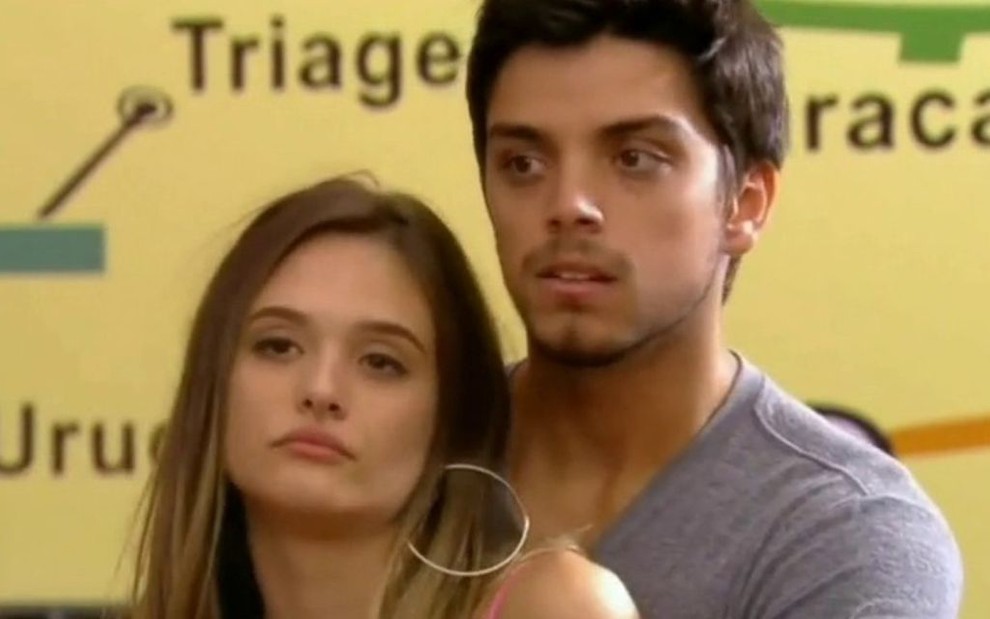 Rodrigo Simas abraça Juliana Paiva por trás; ela está com expressão de confronto em cena de Malhação