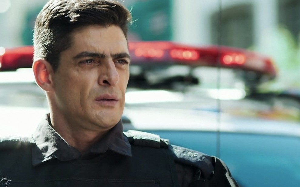 O ator Julio Machado caracterizado como o major Marco Rodrigo, fardado, em frente a uma viatura da polícia em cena de Malhação: Toda Forma de Amor