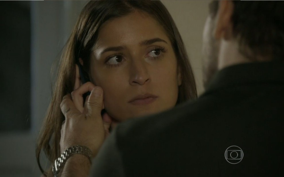 Maria Joana grava com celular na orelha e expressão séria ao lado de Marcelo Faria, de costas na foto