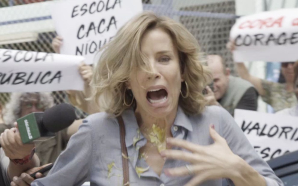 A personagem Malu (Daniela Galli) é atingida por um ovo e faz cara de nojo e susto em cena de Malhação - Viva a Diferença, da Globo; cartazes ao fundo da cena tem escritos de palavras de ordem