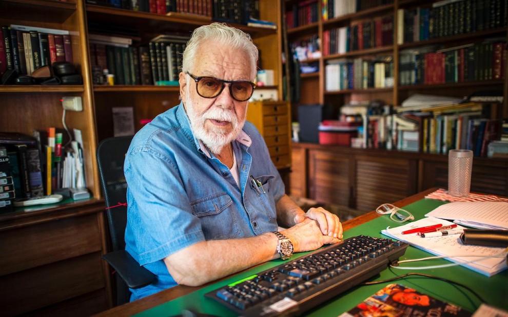 Com uma camisa azul e usando óculos, o autor Manoel Carlos posa em seu escritório sentado em uma mesa diante de um teclado