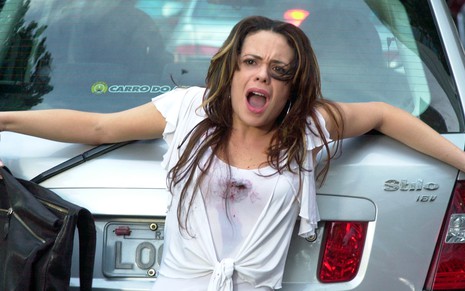 A atriz Vanessa Gerbelli como Fernanda se apoia em um carro após levar um tiro, a sua roupa está suja de sangue na altura do peito, em cena de Mulheres Apaixonadas