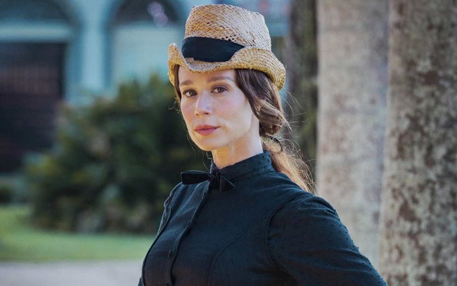 A atriz Mariana Ximenes, com roupa de época, chapéu e vestido preto, em cena como Luísa