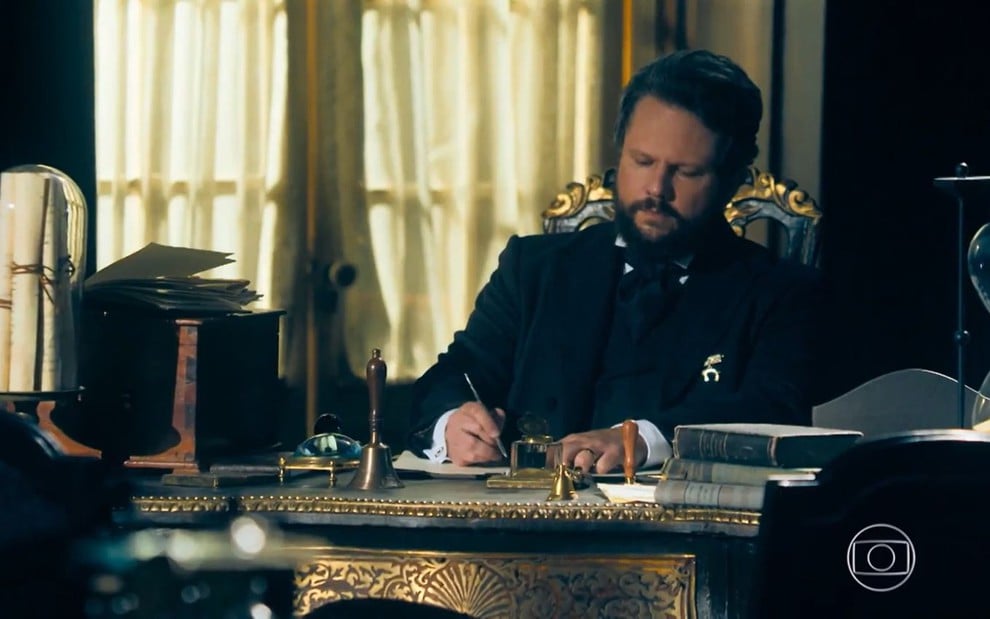 Selton Mello, caracterizado como dom Pedro 2º, sentado à sua mesa em cena da novela Nos Tempos do Imperador
