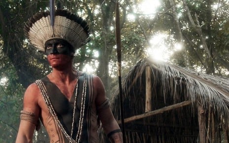 O ator Allan Souza Lima com pintura corporal vermelha e preta e um cocar de penas brancas caracterizado como o índio Ubirajara em cena de Novo Mundo