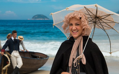A atriz Bia Guedes em uma praia com um bote ao fundo, segura uma sombrinha aberta como Lurdes de Novo Mundo