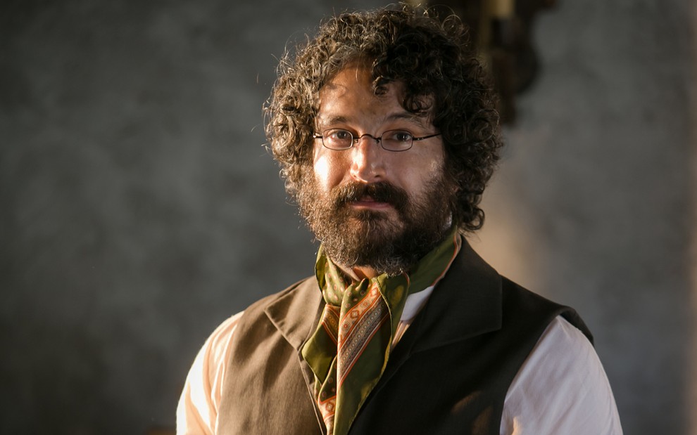 O ator Caco Ciocler com cabelos e barba desgrenhados, de óculos de grau redondos, caracterizado como Peter em cena de Novo Mundo