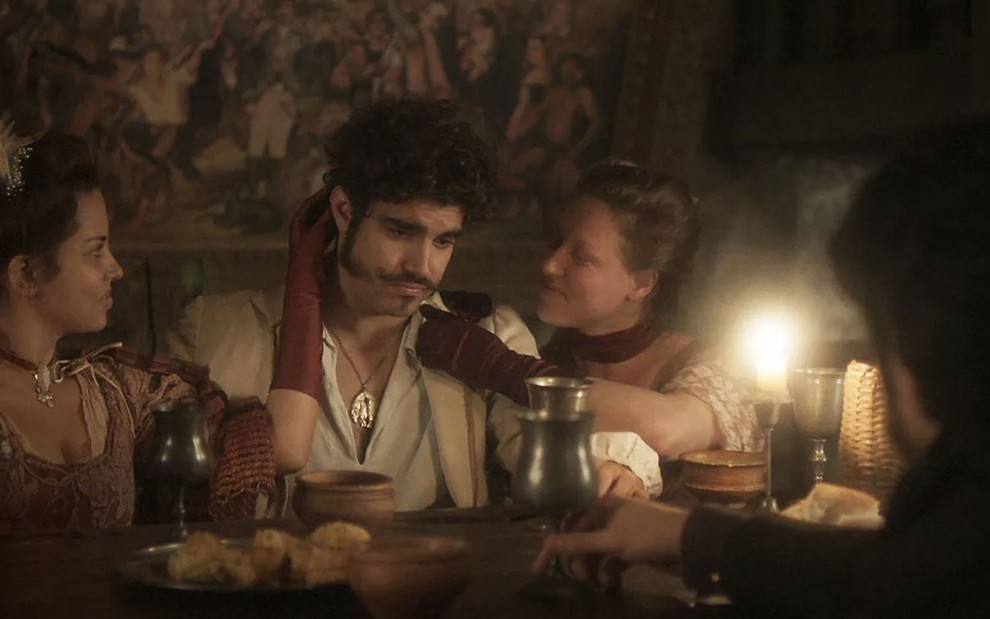 O ator Caio Castro ao centro, com expressão tristonha, agarrado por duas mulheres em uma mesa cheia de bebidas em Novo Mundo
