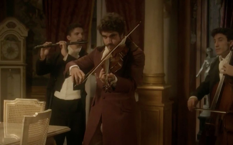 Caio Castro caracterizado como Pedro toca um violino, ao fundo um músico com uma flauta e outro com um violoncelo, em Novo Mundo
