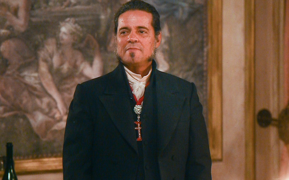 O ator Felipe Camargo está de pé em frente a um quadro caracterizado como José Bonifácio em cena de Novo Mundo