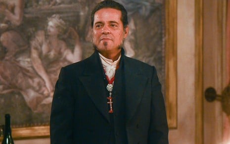 O ator Felipe Camargo está de pé em frente a um quadro caracterizado como José Bonifácio em cena de Novo Mundo