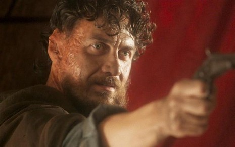 O ator Gabriel Braga Nunes com uma maquiagem especial de queimaduras de terceiro grau no lado direito do rosto, sem orelha, e segurando uma arma em cena de Novo Mundo
