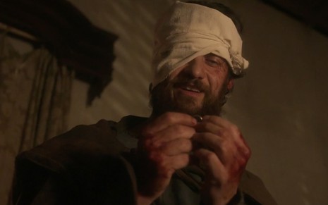 O ator Gabriel Braga Nunes segura um anel com as mãos cheias de sangue e uma bandagem cobrindo parte de sua cabeça como o Thomas em cena de Novo Mundo