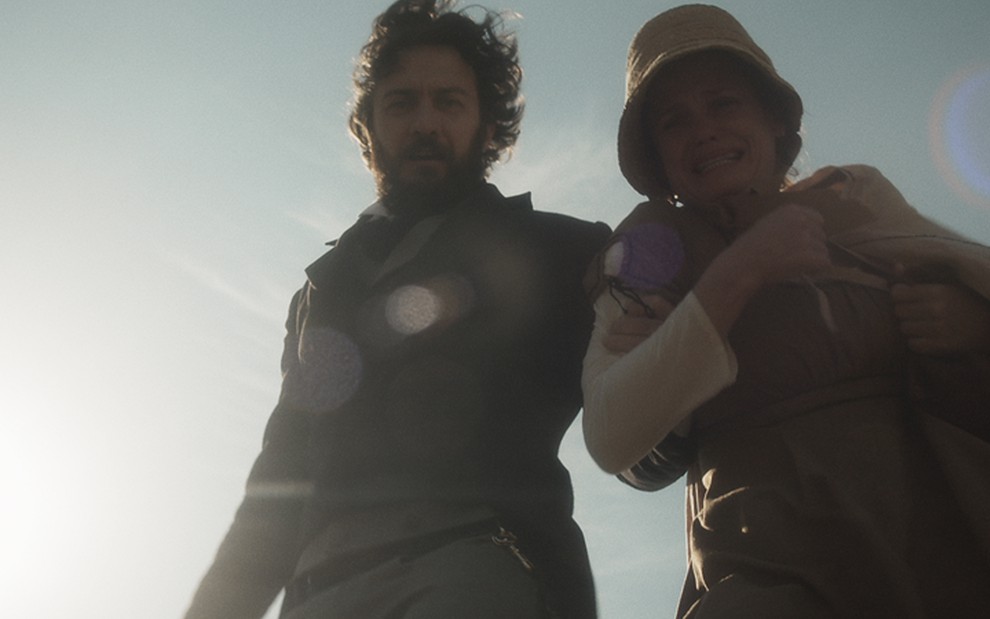 Contra a luz do sol, o ator Gabriel Braga Nunes como Thomas segura a atriz Viétia Zangrandi, a Nívea, pelo pescoço à beira de um penhasco em cena de Novo Mundo