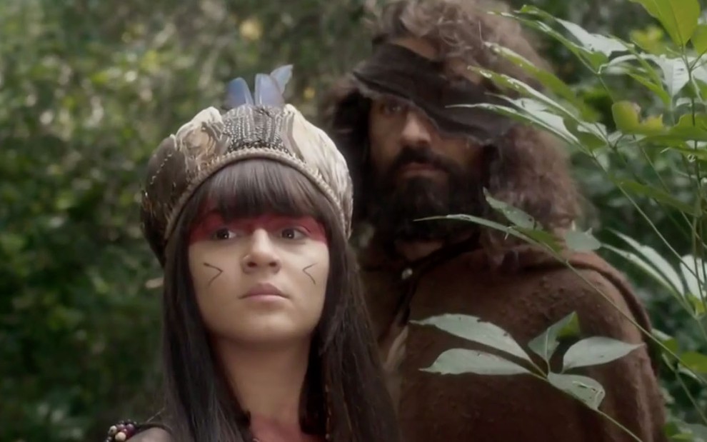 A atriz Giullia Buscacio, a Jacira, com pintura e cocar indígena na frente de Ricardo Pereira, com um pano marrom a cobrir o olho direito, em cena de Novo Mundo