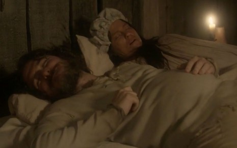 O ator Guilherme Piva, caracterizado como Licurgo, sorri enquanto está deitado em uma cama ao lado de Vivianne Pasmanter, a Germana, em cena de Novo Mundo