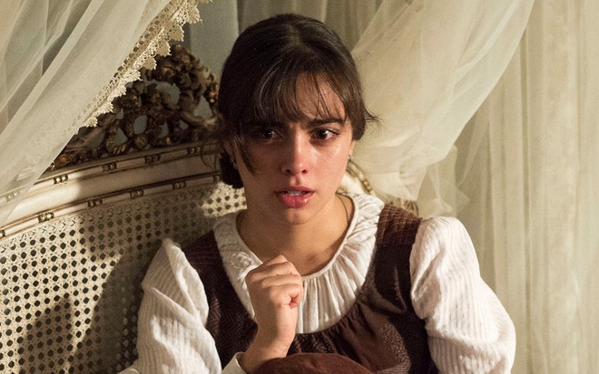 A atriz Isabella Dragão, caracterizada como a Cecília, sentada em uma cama com os olhos vermelhos de chorar em cena de Novo Mundo