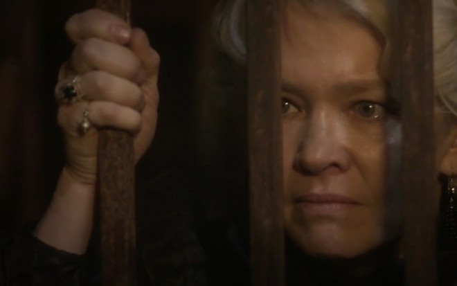 A atriz Julian Lemmertz em um close em seu rosto segura as barras de uma grade com expressão de ódio caracterizada como Greta em cena de Novo Mundo