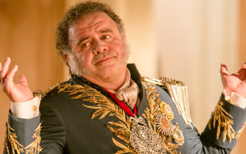 O ator Leo Jaime com trajes históricos, caracterizado como dom João 6º, ergue as mãos em cena da novela Novo Mundo