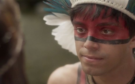 O ator Rodrigo Simas caracterizado como o índio Piatã, com o rosto pintado de vermelho e cocar de penas brancas, em cena de Novo Mundo