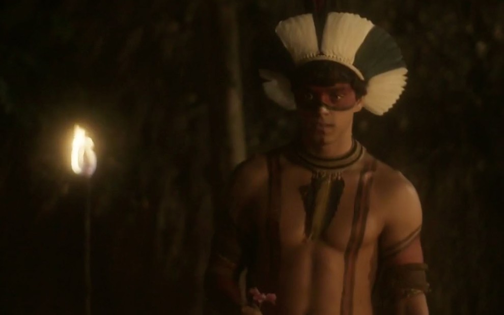O ator Rodrigo Simas com o torso nu e pintura corporal preta e vermelha, com um cocar azul, caracterizado como o índio Piatã de Novo Mundo