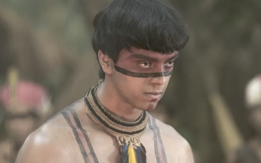 O ator Rodrigo Simas com uma pintura indígena, feita com urucum vermelha nos olhos e tinta preta no corpo, caracterizado como o PIatã em cena de Novo Mundo