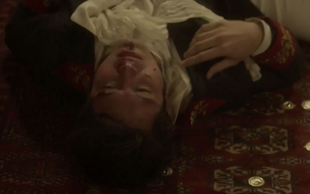 O ator Romulo Estrela, com o rosto bastante machucado e com sangue escorrendo, deitado em um tapete vermelho com moedas de ouro caracterizado como o Chalaça em cena de Novo Mundo