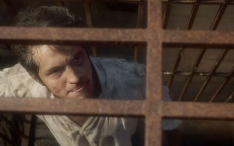 O ator Romulo Estrela com o rosto muito machucado dentro de uma jaula de ferro caracterizado como o Chalaça de Novo Mundo