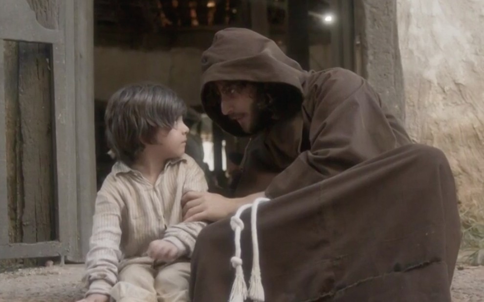 O ator Theo de Almeida Lopes caracterizado como Quinzinho sentado ao lado de Chay Suede, o Joaquim, disfarçado de monge em cena de Novo Mundo