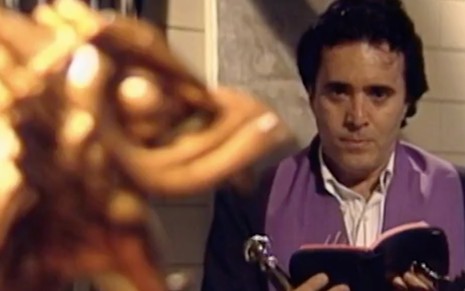 O ator Tony Ramos vestido de padre em frente a uma estátua de dragão dourado com a bíblia aberta em cena de Olho por Olho