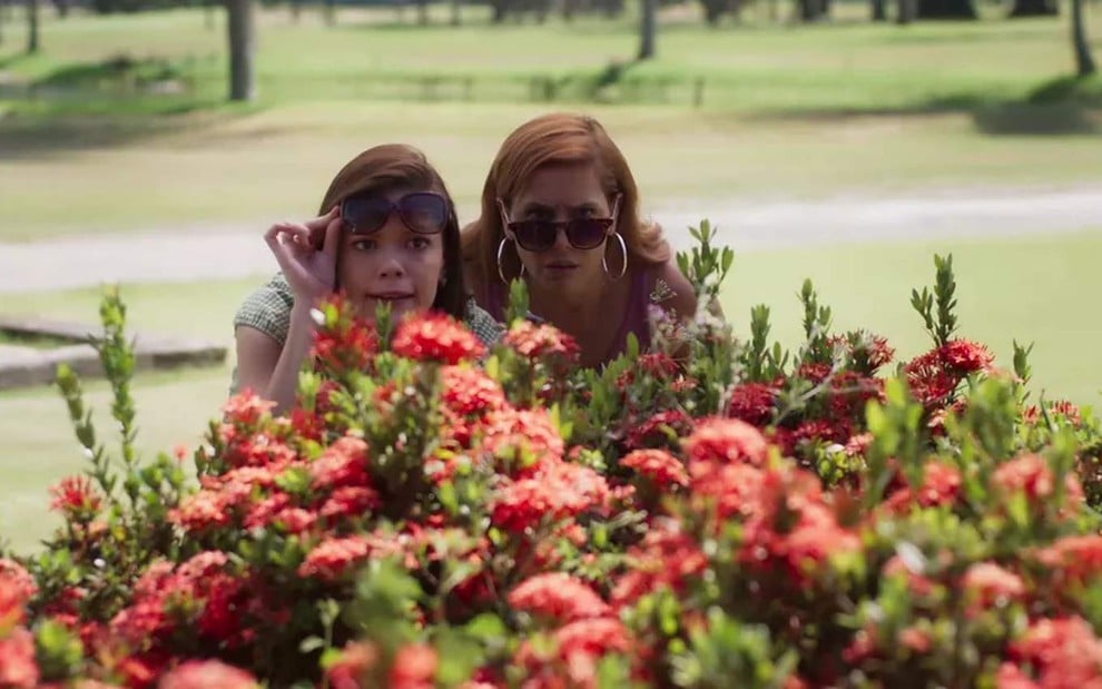 As atrizes Deborah Secco e Vitória Strada escondidas atrás de um arbusto, em cena como Kyra e Alexia em Salve-se Quem Puder