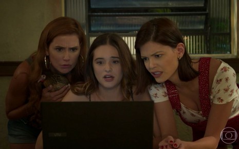 As atrizes Deborah Secco, Juliana Paiva e Vitória Strada em cena de Salve-se Quem Puder: o trio está sentado e olha para uma tela de computador