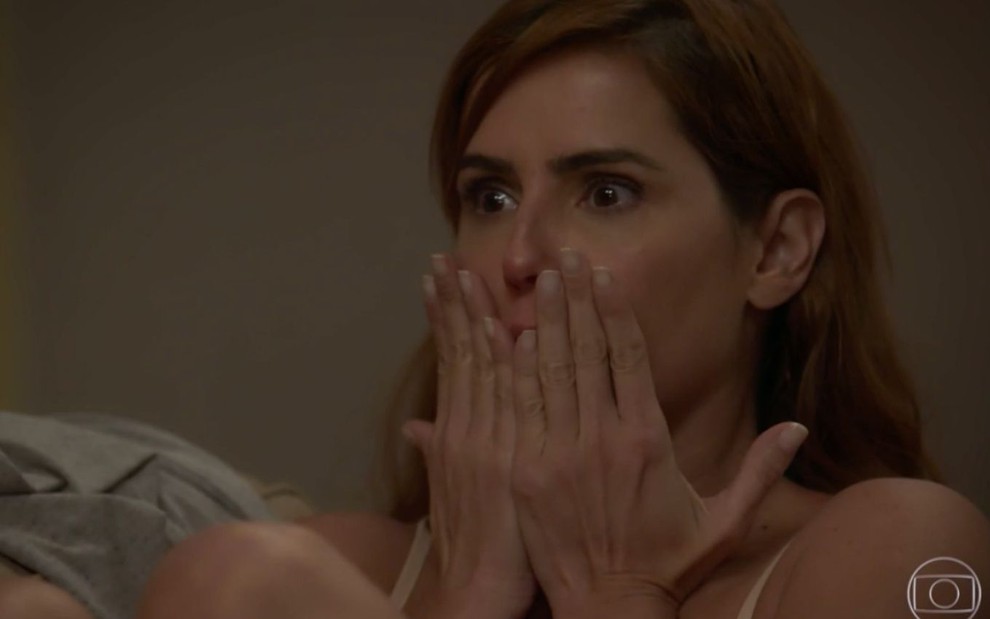 Deborah Secco em cena de Salve-se Quem Puder; caracterizada como Alexia, atriz olha as mãos na boca em sinal de surpresa