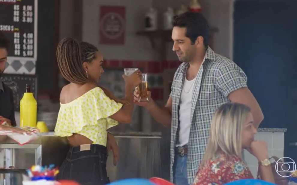 Dandara Mariana grava de blusa amarela com copo de cerveja na mão ao lado de João Baldasserini, de camisa xadrez