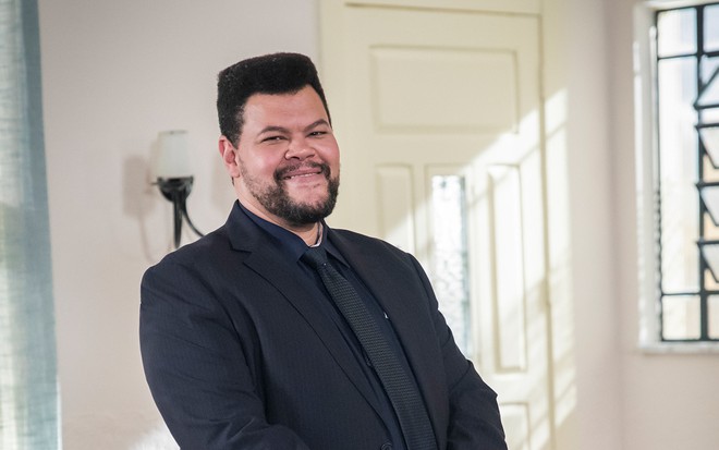 O ator Babu Santana está na sala de uma casa com um terno completamente preto como o policial federal Nanico