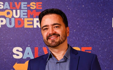 O autor Daniel Ortiz com um blazer preto e uma camisa azul marinho em frente a um backdrop azul com o logo da novela Salve-se Quem Puder