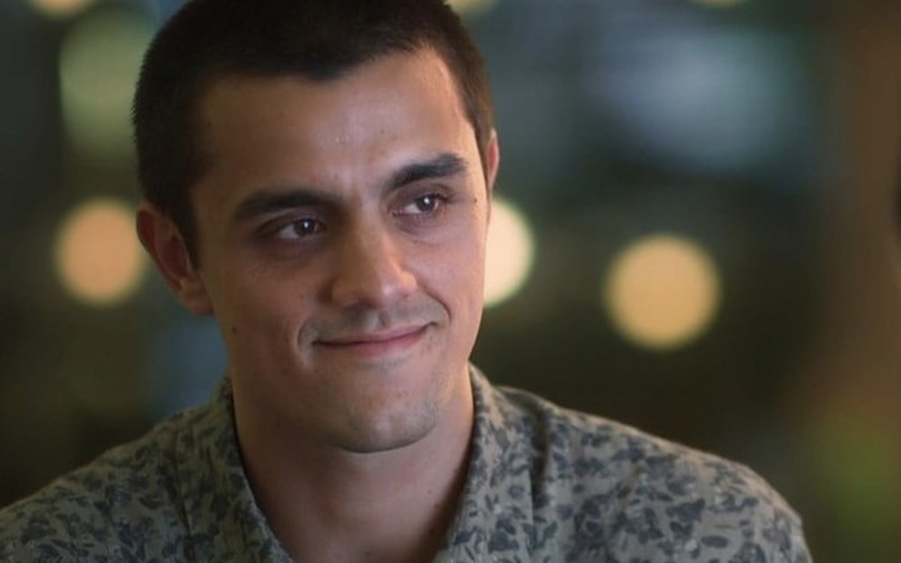 O ator Felipe Simas sorri caracterizado como Téo em frente a um fundo de várias luzes difusas em cena de Salve-se Quem Puder