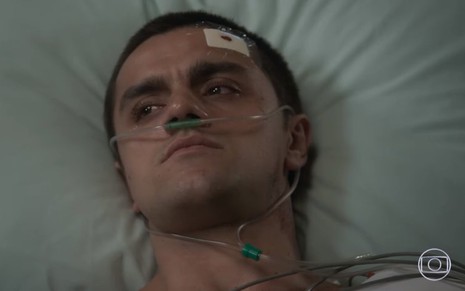 O ator Felipe Simas está deitado em uma cama de hospital, com curativos no rosto, em cena como Téo em Salve-se Quem Puder