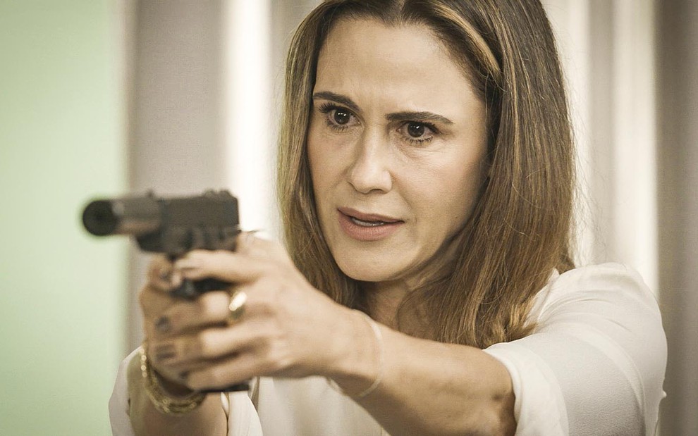 Com expressão de raiva, Guilhermina Guinle segura um revólver com as duas mãos e aponta para a esquerda em cena de Salve-se Quem Puder