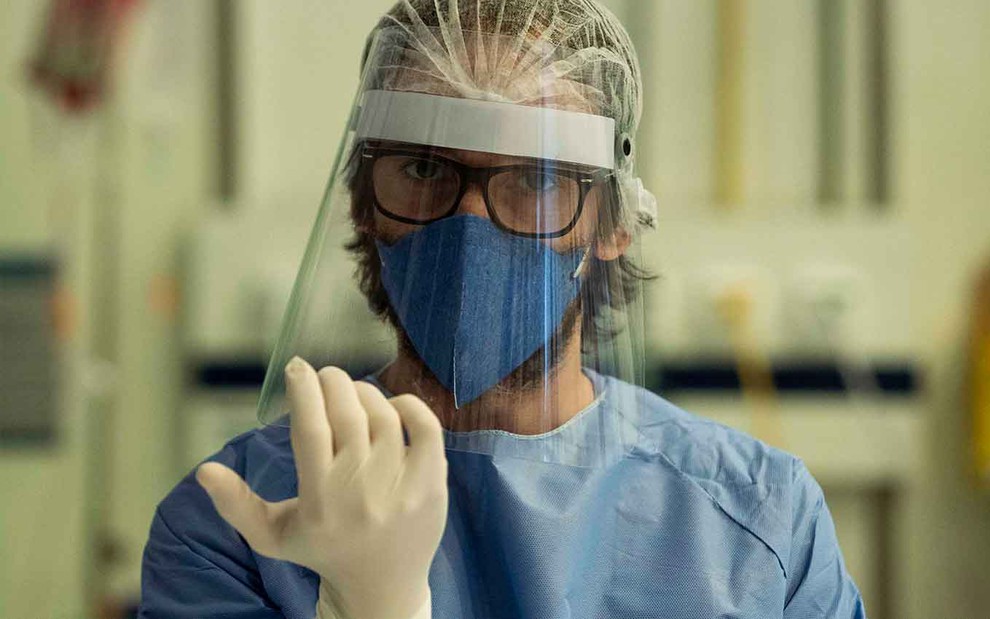 Julio Andrade está com equipamento completo de proteção contra o coronavírus com máscara, luvas, face shield e macacão caracterizado como o médico Evandro em Sob Pressão