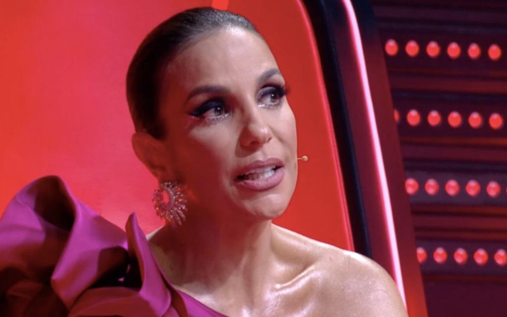 Ivete Sangalo, com um vestido rosa, está sentada em sua cadeira do The Voice e faz cara de concentração