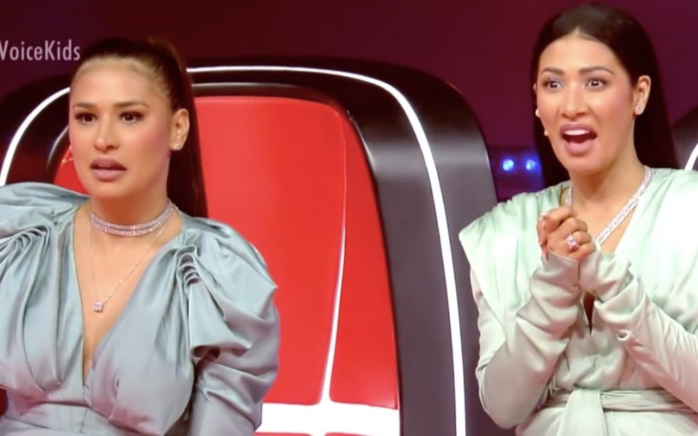 As cantoras Simone e Simaria, ambas de boca aberta, diante de suas cadeiras vermelhas no The Voice Kids